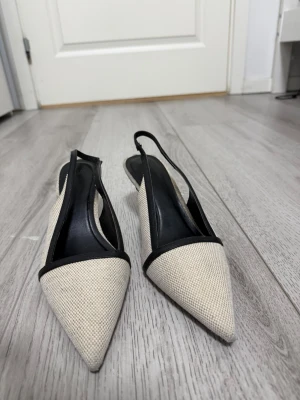 Beige och svarta slingback-pumps H&M - Säljer ett par stilrena pumps från H&M i beige textil med svarta detaljer. Skorna har spetsig tå och öppen häl med slingback-rem. Perfekta för dig som gillar en chic och modern look. Materialet är textil med syntetisk innersula.