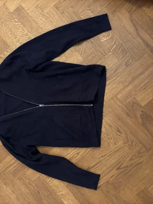 Svart zip-kofta med fickor - En enkel svart kofta med dragkedja framtill och två stora fickor. Koftan har lång ärm och rak passform, perfekt för att slänga på sig när det är lite kyligt. Materialet ser ut att vara mjukt och bekvämt.