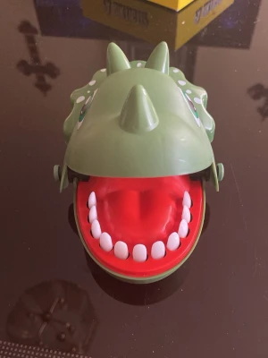 Grön dinosaurie-leksak med tänder - Rolig leksak formad som en grön dinosaurie med öppet gap och vita plasttänder. Leksaken har detaljer som horn och prickar på huvudet, och insidan är röd. Perfekt för spel eller som dekoration.