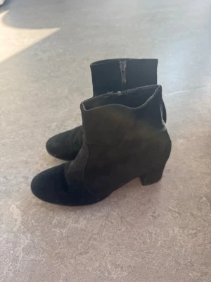 Svarta mockastövletter med klack - Säljer ett par svarta stövletter i mocka-look med rund tå och medelstor klack. Dragkedja på insidan för enkel på- och avtagning. Stövletterna har en stilren och klassisk design som passar till många olika outfits.