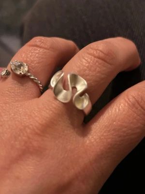 Silvrig ring med vågig design -  silvrig ring med en vågig, abstrakt form som ger ett modernt och konstnärligt intryck. Ringen är tillverkad i metall med matt finish och har en öppen design som gör den justerbar. Perfekt för dig som gillar statement-smycken och vill sticka ut.