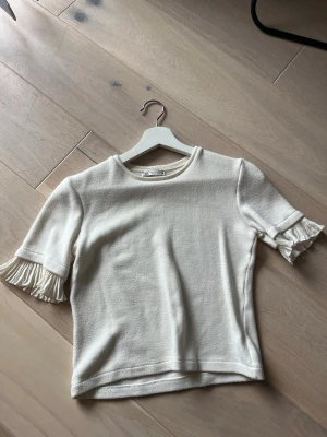Vit topp med volangärm från Zara - Snygg vit topp från Zara med korta ärmar och volangdetaljer vid ärmsluten. Tillverkad i ett mjukt bomullsliknande material med rund halsringning. Perfekt för dig som gillar stilrena plagg med en twist.