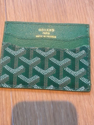 Grön kortfodral från G - Snyggt grönt kortfodral från G med klassiskt mönster i vitt och brunt. Tillverkad i tåligt läder med flera kortfack och guldfärgad text. Perfekt för dig som vill ha något stilrent och exklusivt i fickan.