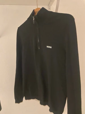 Svart halvzip tröja från BOSS - Snygg svart långärmad tröja från BOSS med halvzip och diskret logga på bröstet. Tröjan har hög krage och är tillverkad i ett mjukt material som passar perfekt till en stilren look.
