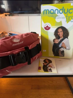 Manduca bärsele i röd bomull - Säljer en Manduca bärsele i röd färg med svart spänne och bred midjerem. Tillverkad i ekologisk bomull och kommer med originalkartong och instruktionshäfte. Perfekt för att bära små barn nära kroppen på ett bekvämt sätt.