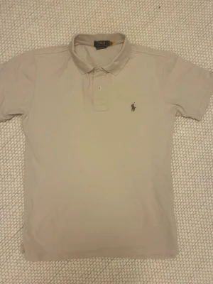 Beige pikétröja från Polo Ralph Lauren - Klassisk beige pikétröja från Polo Ralph Lauren med korta ärmar och broderad logga på bröstet. Tröjan har krage och knäppning med två knappar. Perfekt för en stilren och avslappnad look.