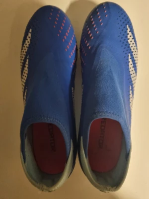 Blå Adidas Predator fotbollsskor - Säljer ett par blå Adidas Predator fotbollsskor med vita och röda detaljer. Skorna har en strumpliknande design utan snörning och är tillverkade i syntetmaterial med High Definition Texture för bättre bollkontroll. Perfekta för dig som vill ha grym touch på planen.