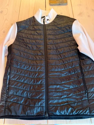 Svart quiltad jacka från J.Lindeberg - Snygg svart quiltad jacka från J.Lindeberg med vita ärmar och hög krage. Jackan har dragkedja framtill och sidofickor med dragkedja. Perfekt för dig som gillar sportig och stilren look. Perfekt till golf eller träning eller till vardags. Pytte liten fläck se sista bilden