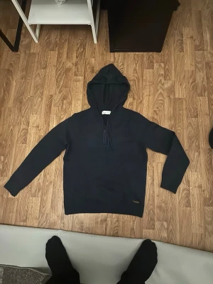 Mörkblå stickad hoodie - Mörkblå stickad hoodie med luva och dragsko. Tröjan har långa ärmar och ribbade muddar vid ärmslut och nederkant. Perfekt för dig som vill ha en enkel och stilren stickad tröja med hoodie-look.
