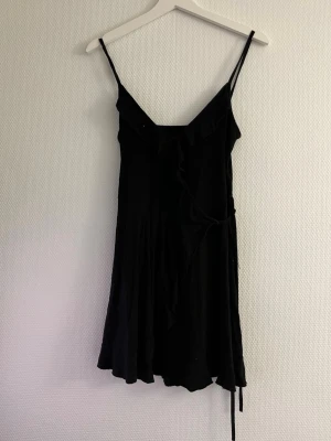 Svart omlottklänning med volang - Snygg svart kortklänning med tunna axelband och omlottmodell. Klänningen har volangdetaljer längs urringningen och knyt i midjan för en feminin siluett. Perfekt för dig som gillar enkel men stilren design.