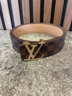 Brunt rutigt bälte i skinn - Snyggt brunt bälte med klassiskt rutigt mönster.  Tillverkat i skinn och designat i Paris. Perfekt accessoar för att lyfta vilken outfit som helst.