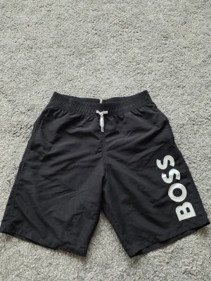 Svarta badbyxor från BOSS - Svarta badbyxor från BOSS med vit logga på ena benet och vit dragsko i midjan. Modellen har elastisk midja och meshfoder på insidan för extra komfort. Perfekta för stranden eller poolen.