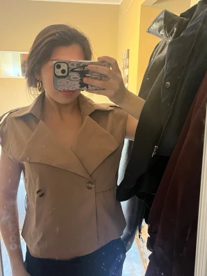 Beige ärmlös dunväst med bälte - Säljer en beige ärmlös dunväst med bred krage och dubbelknäppning framtill. Västen har en tillhörande bälte  som är väldigt snygg till. 