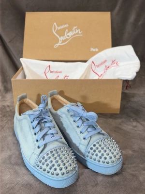 Ljusblå Christian Louboutin sneakers - Snygga ljusblå sneakers från Christian Louboutin i mocka med matchande blå skosnören och coola silverfärgade nitar på tån. Skorna har en klassisk låg siluett och den ikoniska röda sulan. Perfekt för dig som vill sticka ut med stil.