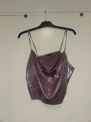 Skimrig lila linnetopp - Säljer en glittrig lila linnetopp med tunna axelband och draperad urringning. Toppen har en croppad passform och skimrar snyggt i ljuset, perfekt för att sticka ut. Materialet känns mjukt och följsamt mot huden.