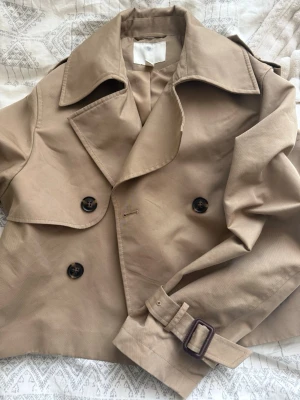 Kort beige trenchcoat med bälte - Stilren kort trenchcoat i beige med dubbelknäppning och stora svarta knappar. Jackan har bred krage, axelklaffar och bälte vid ärmsluten för extra detalj. Perfekt att slänga över en hoodie eller t-shirt för en chill look.