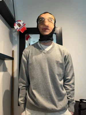 Grå V-ringad tröja från GANT - Klassisk grå V-ringad tröja från GANT med diskret logga broderad på bröstet. Tröjan har långa ärmar, ribbade muddar och är tillverkad i ett mjukt material som passar perfekt till lager-på-lager-stil.