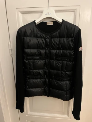 Moncler cardigan - Säljer denna dam Moncler cardigan i storlek S. Modellen har en vadderad front fylld med dun samt ärmar i merinoull. Plagget har knäppning framtill, fickor och den klassiska Moncler-loggan på ärmen. Det är en mångsidig cardigan som kan användas året runt och fungerar lika bra som kofta som en lättare jacka.  Cardiganen är i mycket fint skick och har endast använts ett fåtal gånger, utan några defekter. Äkthetsbevis finns på insidan via QR-tag. Nypris var 11 000 kr. Galge medföljer vid köp. 