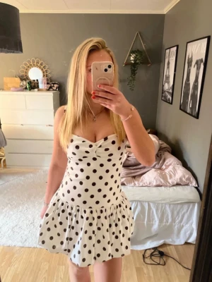 Vit prickig kort klänning - Supersöt vit klänning med svarta prickar och breda axelband. Klänningen har en söt rosettdetalj vid bysten och en volangig kjol som ger en lekfull look. Perfekt för varma dagar och somriga tillfällen.