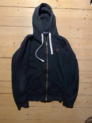 Svart hoodie från Polo Ralph Lauren - Svart hoodie med dragkedja från Polo Ralph Lauren. Klassisk design med vit dragsko i huvan, kängurufickor och röd broderad logga på bröstet. Tillverkad i mjukt bomullsmaterial. Storlek M och passar true to size. Har lite prickar på ena ärmen men märks inte.