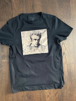 Limitato T-shirt  - Svart t-shirt från Limitato med ett stort konstnärligt motiv på bröstet i beige och svart. T-shirten har rund hals och korta ärmar, perfekt för dig som gillar unika prints och streetstyle. Materialet är mjukt bomull.