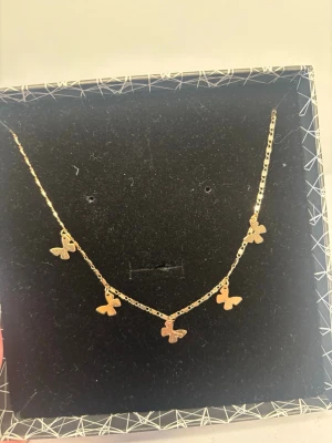 Guldigt halsband med fjärilar - Snyggt halsband i guldfärgad metall med fem små fjärilsberlocker. Kedjan är tunn och smidig.  Perfekt accessoar för att lyfta vilken outfit som helst.
