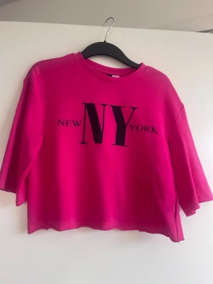 Rosa croppad t-shirt i oversize modell - Snygg rosa croppad t-shirt från H&M med trycket 'NEW YORK' i svart framtill. T-shirten har rund halsringning och lite vidare, korta ärmar. Perfekt statement-plagg för dig som gillar färg och cityvibes. Helt ny med prislapp och är i oversize modell. Skulle passa de flesta storlekar.