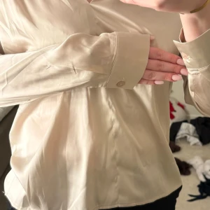 Beige blus med v-ringning - Snygg beige blus med v-ringning och långa ärmar. Blusen har knappar vid ärmsluten och är tillverkad i ett tunt, lätt material som ger en elegant känsla. Perfekt att matcha med jeans eller kjol för en stilren look.