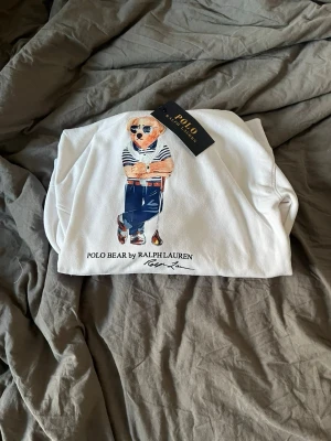 Vit hoodie Polo Bear Ralph Lauren - Vit hoodie från Ralph Lauren med ikoniskt Polo Bear-motiv på framsidan. Klassisk huva och ribbade muddar. Materialet är mjukt och bekvämt, perfekt för en chill look. Snyggt tryck med björnen i jeans och randig tröja. Taggen sitter kvar.