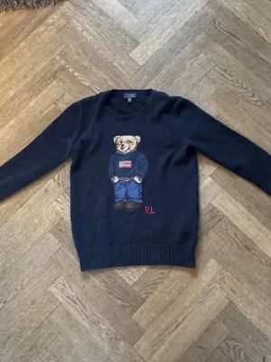 Polo Bear sweatshirt RL - Säljer en mörkblå stickad tröja från Polo Ralph Lauren med ikoniskt björnmotiv på bröstet. Tröjan har rund halsringning och ribbstickade muddar. Björnen bär jeans och en tröja med amerikansk flagga, samt RL-broderi i rött.