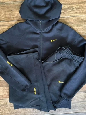 Svarta NOCTA mjukisbyxor från Nike - Säljer ett par svarta mjukisbyxor från Nike NOCTA-kollektionen. Byxorna har gul NOCTA-logga på benet och gul Nike Swoosh framtill. Tillverkade i mjukt material med resår i midjan och dragsko för skön passform. Perfekta för chill eller streetwear.