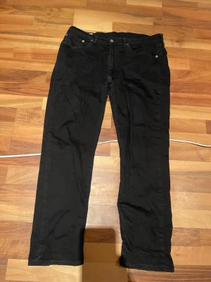 Svarta raka jeans från Levi's - Säljer ett par klassiska svarta jeans från Levi's med rak passform och fem fickor. Jeansen har en enkel design med svarta sömmar och metallknappar. Perfekta för dig som gillar en clean och tidlös look.