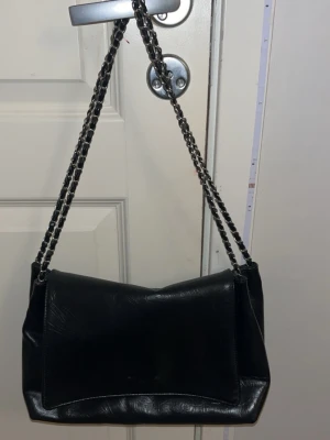Svart axelväska med kedjerem - Snygg svart axelväska i slätt konstläder med dubbla kedjeremmar i silverfärg. Väskan har en enkel, stilren design med lock och rektangulär form. Perfekt för dig som gillar en klassisk men edgy look.