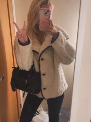Krämvit teddyjacka med svarta detaljer - Säljer en fluffig och trendig krämvit teddyjacka med stora krage och svarta kontrastsömmar. Jackan har knäppning framtill och en boxig, avslappnad passform. Perfekt för dig som vill ha en mysig och snygg look nu i vår utan att den är för kall eller varm❣️bara använd några få gånger 