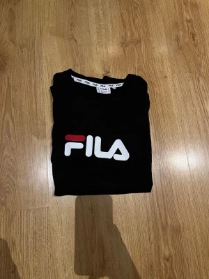 Svart FILA t-shirt med logga - Snygg svart t-shirt från FILA med stor vit och röd logga på bröstet. Klassisk rund halsringning och mjukt material som känns skönt mot huden. Perfekt för dig som gillar sportiga och stilrena plagg med tydlig branding.