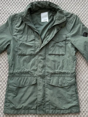 J.Lindeberg fieldjacket - Stilren grön vindjacka från J.Lindeberg med fyra stora fickor framtill och hög krage. Jackan har dragkedja och tryckknappar, samt en patch på ena ärmen. Perfekt för dig som gillar en clean och funktionell look.