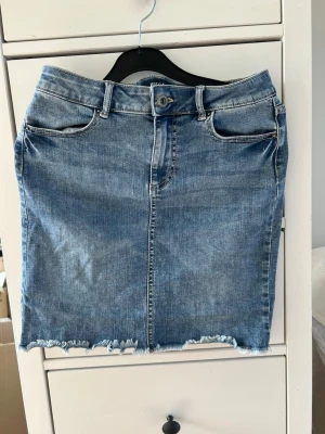 Blå jeanskjol - Säljer en klassisk blå jeanskjol med rå, fransig kant nertill. Kjolen har fem fickor, bälteshällor och stängs med knapp och dragkedja framtill. Perfekt för dig som gillar en trendig stil i vår och sommar💗