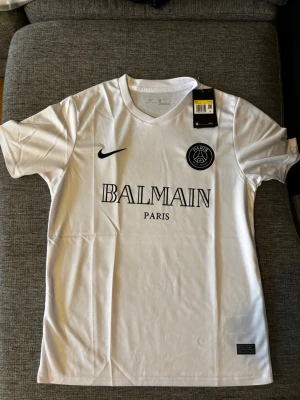 Balmain x PSG fotbollströja - Snygg vit t-shirt från Nike i samarbete med PSG och Balmain. Tröjan har rund hals, svart Nike-logga, PSG-märke och Balmain Paris-tryck på bröstet. Tillverkad i lätt och ventilerande Dri-FIT-material, perfekt för träning eller streetwear.