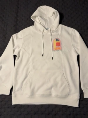 Vit hoodie från Casablanca - Säljer en vit hoodie från Casablanca med färgglad grafisk print fram och stort tryck på ryggen. Hoodien har huva med dragsko och en stor magficka. Materialet känns mjukt och skönt, perfekt för en chill och stilren look. STORLEK M, PASSAR S-M. HELT NY!