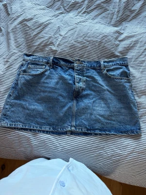 Blå jeanskjol i klassisk modell - Säljer en blå jeanskjol i klassisk femficksmodell med dragkedja och knapp framtill. Kjolen är kort och har en rak passform, perfekt till vardagslooken. Märket är 157 och materialet är slitstark denim.