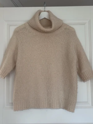 Beige stickad polotröja - Mysig beige stickad polotröja med halvlånga ärmar och bred polokrage. Tröjan har en avslappnad passform och är gjord i ett fluffigt, mjukt material som känns lyxigt. Perfekt för lager på lager och en stilren look.