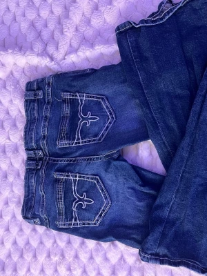 Mörkblå bootcut jeans med brodyr - Säljer ett par mörkblå bootcut jeans med slitningar och snygg vit brodyr på bakfickorna. Jeansen har låg midja och kontrastsömmar som ger en cool look. Perfekta för dig som gillar en avslappnad men trendig stil.