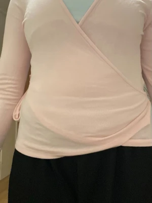 Ljusrosa omlottopp med knyt - Supersöt ljusrosa omlottopp med långa ärmar och knyt i sidan. Toppen har en mjuk och stretchig känsla, perfekt för att skapa en avslappnad men ändå stilsäker look. Passar perfekt till både jeans och kjol.