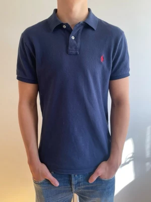 Marinblå Ralph Lauren Pike - Marinblå Ralph Lauren pike med röd broderad logga på börstet. Piken är storlek L men sitter som en M modellen på bilden är 182cm lång👍 Perfekt inför sommaren och våren piken är i helt nytt skick🤩☀️ nypris:1695kr mot mitt pris 449kr😃✅ har du några funderingar så är det bara att skriva💯