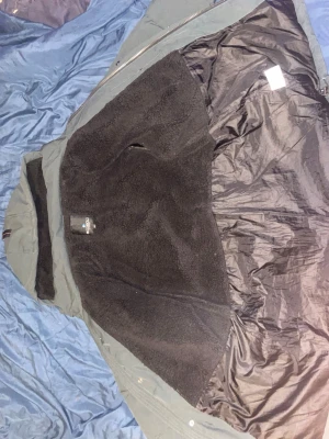 Teddyjacka från Cobolt of Sweden - Mysig teddyjacka från Cobolt of Sweden med svart fluffigt foder och yttertyg i grått. Jackan har dragkedja och huva, perfekt för kalla dagar. Materialet är mjukt och värmande, och designen är enkel men stilren.