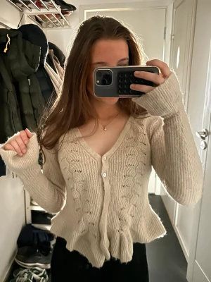 Beige kabelstickad kofta med volang - Söt beige kabelstickad kofta med volangkant nedtill och v-ringning. Koftan har långa ärmar, ribbade muddar och knäpps med vita knappar framtill. Perfekt för dig som gillar en mjuk och feminin stil.