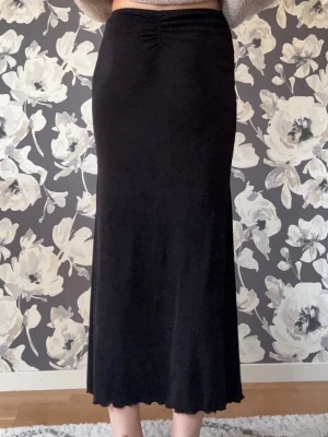 Svart långkjol med vågig kant - Säljer en svart långkjol med enkel och stilren design. Kjolen har en lätt utsvängd passform och vågig kant längst ner för en snygg detalj. Perfekt att styla med crop tops eller stickade tröjor. Jag är 173 vilket gör den lite kort för mig i min smak. Använd ungefär 2 gånger.