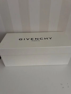 Vit Givenchy kartong - Säljer en stilren vit skokartong från Givenchy Paris. Kartongen är rektangulär med svart logotyp på locket och passar perfekt för förvaring eller som inredningsdetalj. Materialet är hård kartong och designen är minimalistisk, mått 34*15 , hög 13 cm.