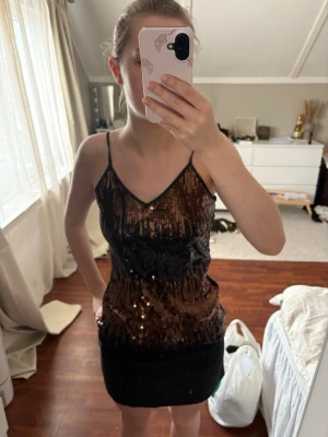 Svart paljettklänning med smala band - Säljer en svart kort klänning med tunna axelband och v-ringning. Framsidan är täckt av glittriga paljetter i svart och brons, medan baksidan är i genomskinligt tyg. Perfekt för dig som vill sticka ut med en festlig look!