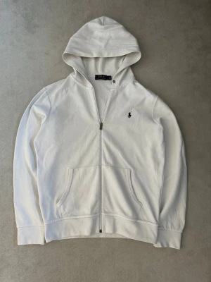 Polo Ralph Lauren - Snygg vit hoodie med dragkedja från Polo Ralph Lauren. Klassisk design med huva, fickor framtill och den ikoniska lilla loggan broderad på bröstet. Tillverkad i mjukt bomullsmaterial som känns skönt mot huden. Perfekt för en avslappnad och stilren look. 8/10 i skick. Är i storlek M men tror även den passar S oxå!
